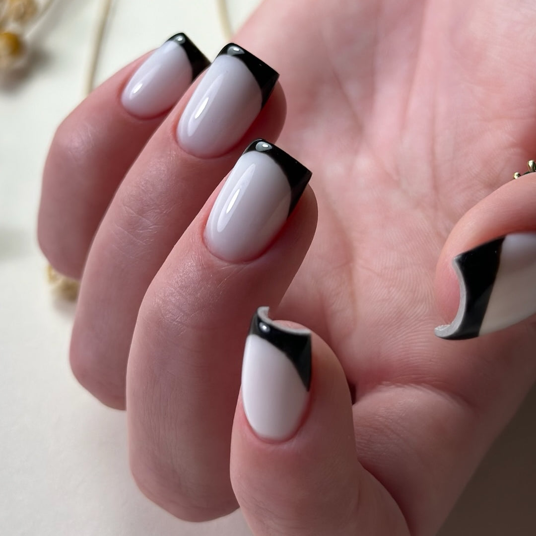 A close-up image of a hand showcasing a manicure crafted with Paloma builder gel, featuring long square nails with a glossy finish. The nails are painted in a classic French tip style, with a modern twist of broad black tips that elegantly contrast with the milky white base. The manicure demonstrates a sophisticated and bold aesthetic, set against a soft-focus background that accentuates the sharpness of the nail art.